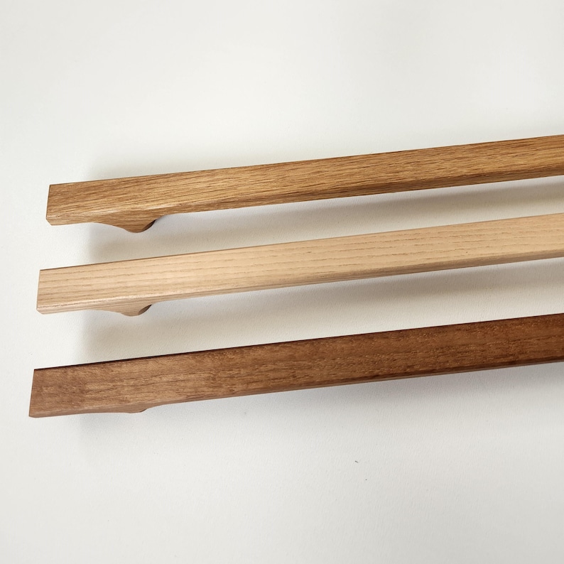 May include: Three wooden poster hangers in varying shades of brown. The top hanger is light brown, the middle is a natural wood color, and the bottom is a dark brown. Each has a curved handle-like design.