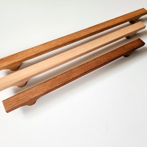 May include: Three wooden cabinet pulls in varying shades of brown. The pulls are rectangular with a curved handle design. The wood tones range from light to dark, showcasing the natural grain.