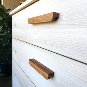 May include: Close-up of a light-colored wooden dresser with three drawers. Each drawer has a horizontal, rectangular wooden handle with a curved edge. The handles are a darker shade of brown than the dresser.