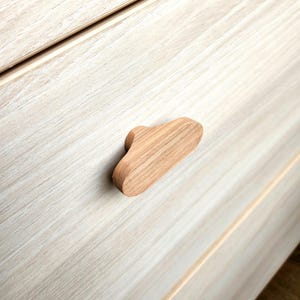 May include: A light-colored wooden cabinet pull with a cloud-like shape. The pull is made of natural wood and is mounted on a light-colored wooden cabinet. The cabinet has a light wood grain pattern.