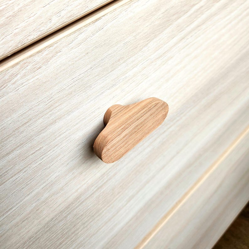 May include: A light-colored wooden cabinet pull with a cloud-like shape. The pull is made of natural wood and is mounted on a light-colored wooden cabinet. The cabinet has a light wood grain pattern.