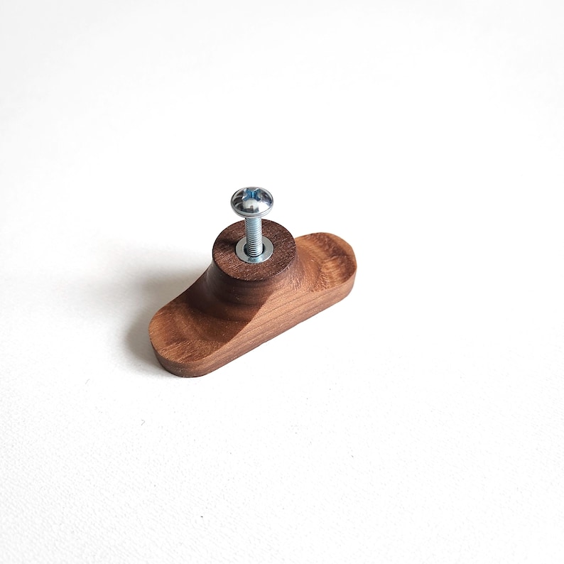 May include: A wooden cabinet knob with a dark brown finish. The knob has a rounded, oblong shape with a cylindrical base. A silver screw is inserted through the center for attachment. The knob is designed for furniture and cabinetry.