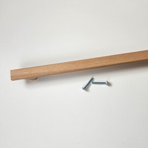 May include: A light brown wooden cabinet pull with a rectangular design. Two silver screws are positioned near the handle. The handle is made of light-colored wood and has a smooth finish.