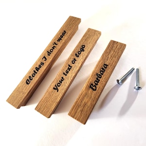 May include: Three wooden drawer pulls with engraved text. The first reads "Clothes I don't wear", the second "Your text or logo", and the third "Barbara". Two silver screws are next to the pulls. The wood has a natural grain.