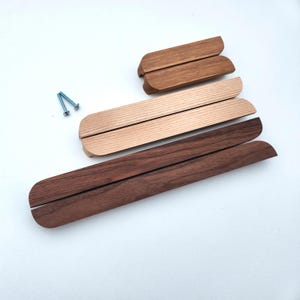 Könnte beinhalten: Eine Sammlung von sechs Holzschrankgriffen in verschiedenen Längen und Holztönen, von hell- bis dunkelbraun, angeordnet in einer Treppenformation. Zwei kleine silberne Metallschrauben befinden sich links neben den hölzernen Beschlagteilen.