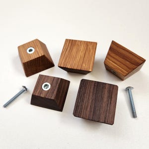 Op de afbeelding: Vijf houten kastknoppen in verschillende vormen en houtnerven. De knoppen zijn in bruintinten en hebben een metalen inzetstuk voor montage. Twee zilveren schroeven zijn ook zichtbaar, wat wijst op een eenvoudige installatie. Deze knoppen zijn ideaal om een natuurlijk tintje aan meubels toe te voegen.