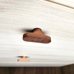 May include: Close-up of a wooden cabinet knob with a unique T-shape design. The knob is made of dark brown wood and is mounted on a light-colored cabinet door. The image highlights the natural wood grain and the knob's smooth, rounded edges.