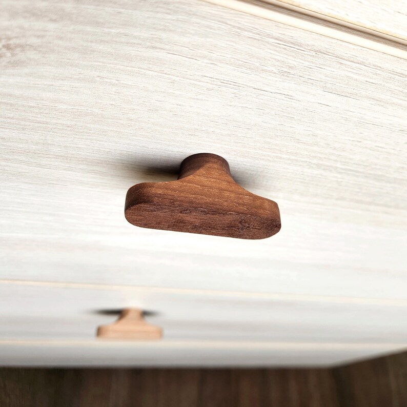 May include: Close-up of a wooden cabinet knob with a unique T-shape design. The knob is made of dark brown wood and is mounted on a light-colored cabinet door. The image highlights the natural wood grain and the knob's smooth, rounded edges.
