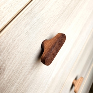 May include: Close-up of a wooden cabinet knob with a rounded, ergonomic design. The knob is a rich, dark brown color, contrasting against the light wood grain of the cabinet. The knob is approximately 3 inches long.
