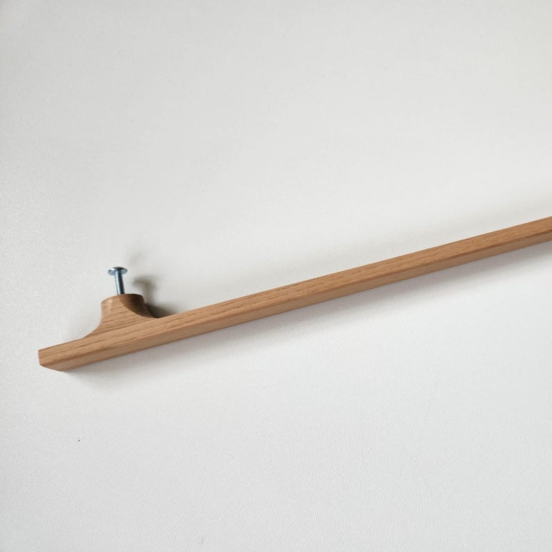 May include: A wooden wall-mounted picture hanger. The hanger is made of light brown wood and has a simple, minimalist design. A single screw secures the hanger to the wall. The hanger is designed to display artwork or photos.