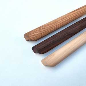 May include: Three wooden trim pieces with a distinct triangular profile. The top piece is light brown with a visible grain, the middle piece is dark brown, and the bottom piece is a pale, smooth wood. Each piece features a flat base and angled sides.