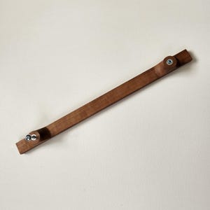 May include: A wooden cabinet pull with a dark brown finish. The rectangular pull has rounded ends with a circular knob and a metal screw on each end. The pull is angled across a neutral background.