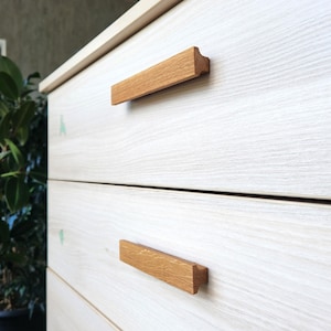 Puede incluir: Primer plano de una cómoda de madera de color claro con tres cajones. Cada cajón tiene un tirador rectangular de madera marrón claro. La veta de la madera es visible en la cómoda y en los tiradores.