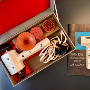 May include: Vintage electric massager kit in a box with accessories. The kit includes a white and red massager, orange attachments, and a booklet with Russian text. The box is open, revealing the contents.
