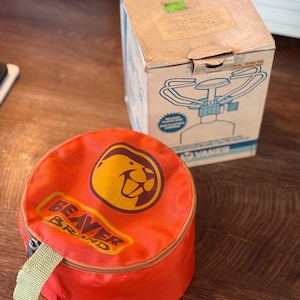 Könnte beinhalten: Eine runde, leuchtend orangefarbene Aufbewahrungstasche mit Reißverschluss, gelbem Biber-Logo und dem Schriftzug BEAVER BRAND, dazu ein rechteckiger Karton mit einer Strichzeichnung eines Campingkochers und dem Aufdruck VANES.