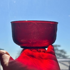 Può includere: Una piccola ciotola di vetro rosso rubino con una superficie strutturata. La ciotola è tenuta contro un cielo blu brillante, con la luce che brilla attraverso il vetro. La forma della ciotola è rotonda con un bordo leggermente svasato.