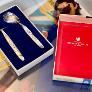Może przedstawiać: Srebrny zestaw łyżki i widelca z ozdobnymi uchwytami, zapakowany w pudełko wyłożone niebieskim aksamitem. Zestaw znajduje się obok czerwonego pudełka z napisem "ORIGINAL EICHHORN-BESTECKE SOLINGEN". Łyżka ma wzór liścia na cząści nabierczej.