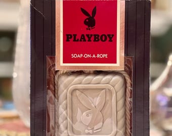 May include: A rectangular box containing a beige soap-on-a-rope with a rope loop. The packaging is black with a red centre panel featuring the Playboy rabbit logo and the text PLAYBOY SOAP-ON-A-ROPE. The soap is embossed with the logo and weighs 180g.