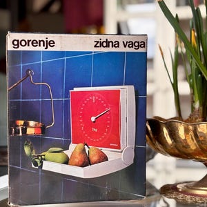 Puede incluir: Caja de báscula de pared Gorenje vintage con esfera roja, que muestra 3 kg. La caja presenta un fondo de baldosas azules y una repisa blanca con peras. El texto "gorenje zidna vaga" está impreso.