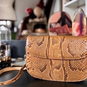 Peut inclure: Un sac à main marron et beige à motif peau de serpent, avec une longue bandoulière réglable. Le sac a une forme incurvée avec une fermeture à rabat. L'arrière-plan comprend des objets décoratifs et des miroirs.