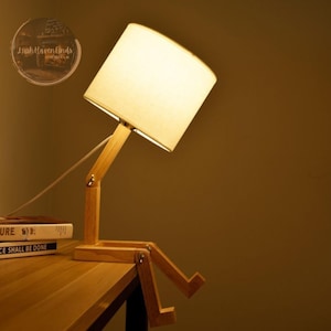May include: A wooden table lamp shaped like a person sitting with its legs crossed. The lamp has a white fabric shade and a wooden base.