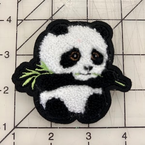May include: Embroidered panda bear patch with black and white fur, brown eyes, and a green bamboo sprig. The patch has a black border and is likely for clothing or accessories.