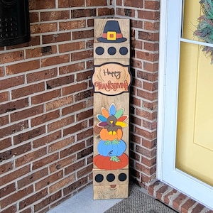 Peut inclure: Panneau en bois sur le th&egrave;me de l'Action de gr&acirc;ce avec un chapeau de p&egrave;lerin, l'inscription "Happy Thanksgiving" et des citrouilles empil&eacute;es. Le panneau est plac&eacute; sur un porche avec une porte jaune et une couronne.