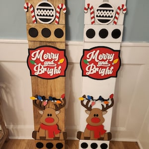 Peut inclure: Deux d&eacute;corations de No&euml;l en bois. L'une est en bois teint&eacute;, l'autre est peinte en blanc. Chacune a un motif de canne en sucre et de boule de No&euml;l en haut, les mots "Merry and Bright", et un renne avec une &eacute;charpe rouge. Le renne a une guirlande lumineuse.