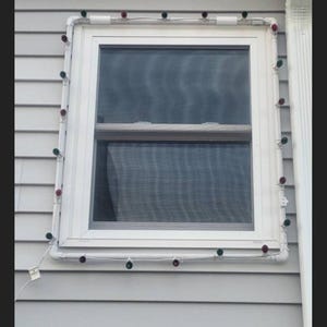 May include: A white window framed with white PVC piping and adorned with Christmas lights. The lights alternate between red and green bulbs. The window is set against gray siding.