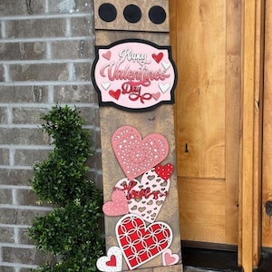 May include: A wooden Valentine's Day sign with a rustic finish. The sign features various heart designs in red, pink, and white, along with the text "Happy Valentine's Day." The sign is decorated with XOXO symbols and black circles.