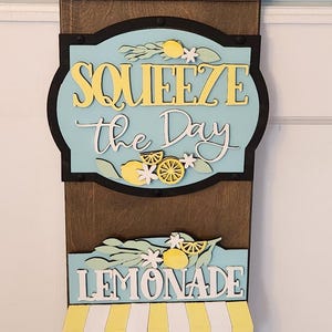 May include: Wooden lemonade stand sign with the text "Squeeze the Day" and "Lemonade". The sign features yellow lemons, flowers, and a basket of lemons. The sign is painted in shades of brown, yellow, and blue.