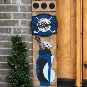 May include: Wooden golf-themed sign with a blue golf cart, a golf ball with crossed clubs, and a golf bag. The sign has the word "Welcome" and three black circles. The sign is made of wood and is painted in blue, black, and white.