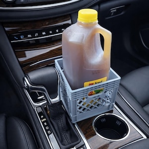 May include: A large plastic jug of iced tea with a yellow cap sits inside a small gray plastic crate in a car's center console. The car interior features wood grain trim and black leather seats. The jug has a yellow label.