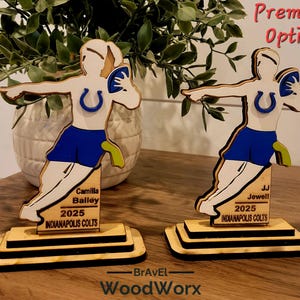 May include: Two wooden Indianapolis Colts football player figurines. Each figure is in a throwing pose, wearing blue shorts and a white jersey with a blue horseshoe logo. The base includes the name, year, and team name. The text "Premium Option" is visible.