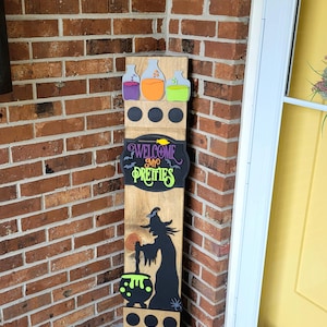 Peut inclure: Panneau en bois d'Halloween avec une silhouette de sorci&egrave;re, un chaudron et le texte "WELCOME MY PRETTIES". Le panneau pr&eacute;sente &eacute;galement des fioles de potion color&eacute;es et des cercles noirs. Il est plac&eacute; pr&egrave;s d'un mur de briques et d'une porte jaune.