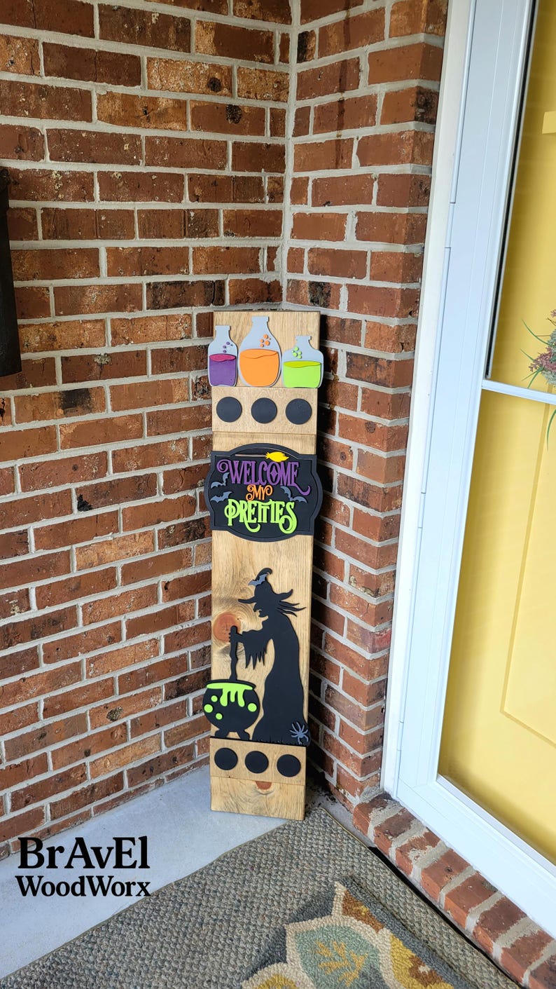 Peut inclure: Panneau en bois d'Halloween avec une silhouette de sorci&egrave;re, un chaudron et le texte "WELCOME MY PRETTIES". Le panneau pr&eacute;sente &eacute;galement des fioles de potion color&eacute;es et des cercles noirs. Il est plac&eacute; pr&egrave;s d'un mur de briques et d'une porte jaune.