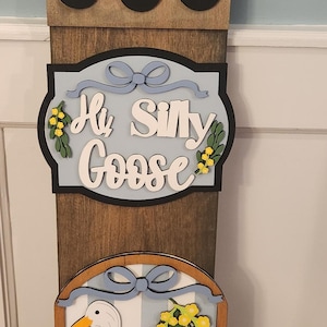 May include: A vertical wooden sign featuring two white goose illustrations, yellow flowers, and light blue bows. The center plaque displays the text "Hi Silly Goose" in white script. The sign includes two rows of three black circular cutouts.