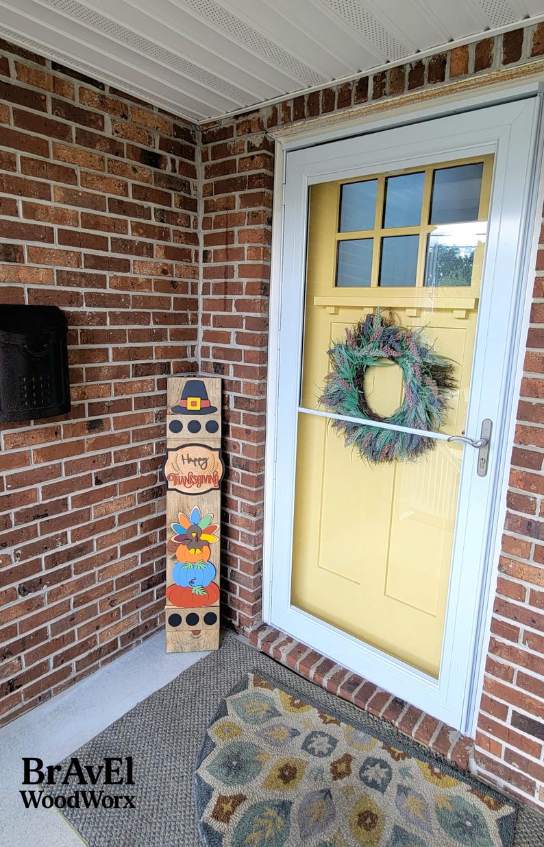 Peut inclure: Panneau en bois sur le th&egrave;me de l'Action de gr&acirc;ce avec un chapeau de p&egrave;lerin, l'inscription "Happy Thanksgiving" et des citrouilles empil&eacute;es. Le panneau est plac&eacute; sur un porche avec une porte jaune et une couronne.