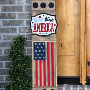 May include: Wooden American flag-themed sign with the text "God Bless America" and a Liberty Bell design. The sign features red and white stripes, blue stars, and black circular accents, perfect for patriotic decor.