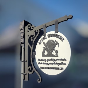 May include: A gray and white sign for Bravel Woodworx, featuring a circular emblem with woodworking tools. The sign includes the text "Building quality products that bring people together" and the website address.
