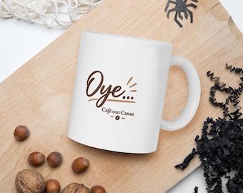 May include: A white ceramic mug featuring the brown text Oye... Café con Canas and a small coffee bean icon. The mug has a standard handle and a smooth finish, displaying the printed design clearly on the front surface of the cylindrical vessel.