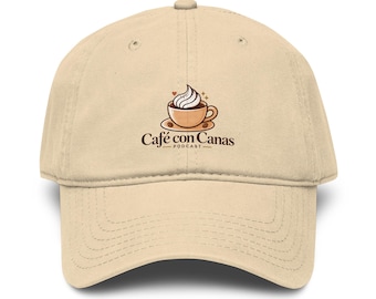 May include: A beige baseball cap featuring a centered graphic of a coffee cup with whipped cream and the text Café con Canas PODCAST. The cap has a structured crown, a curved brim with visible stitching, and a button at the top center.