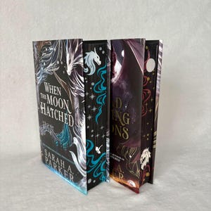 May include: Four fantasy novels with striking covers. The spines display titles such as "When the Moon Hatched" and "Finding Dragons." The covers feature dragon illustrations, celestial elements, and detailed designs in black, teal, and gold. The author's name is visible.