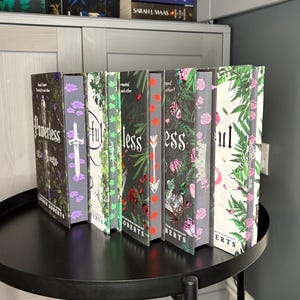 Può includere: Una collezione di libri con copertina rigida con disegni floreali e botanici. I dorsi mostrano titoli come "Powerless" e "Soul" con nomi di autori. I libri sono disposti su un tavolo rotondo nero, con uno sfondo neutro.