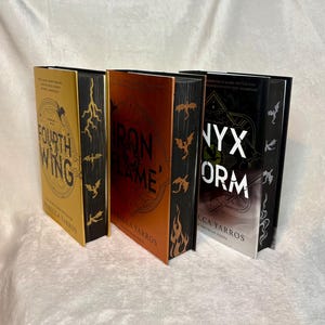 May include: Three hardback books with gold, orange, and black covers. The books are titled "Fourth Wing", "Iron Flame", and "Nyx Storm" by Rebecca Yarros. Each book features dragon and lightning bolt designs.