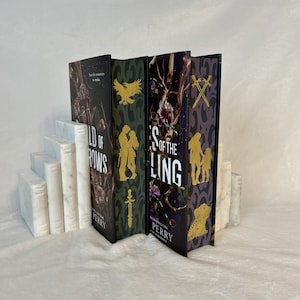 Op de afbeelding: Een verzameling hardcover boeken met decoratieve covers, waaronder titels als "Wild of Rows" en "S of the Ling". De boeken worden geflankeerd door witte marmeren boekensteunen, waardoor een visueel aantrekkelijke presentatie ontstaat. De boekomslagen hebben gouden en donkere kleurenschema's.