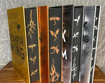 May include: A collection of seven books arranged in a row, featuring metallic gold, copper, and silver spines and covers. The books display various graphic designs including lightning bolts, dragons, flames, and text such as Fourth Wing.