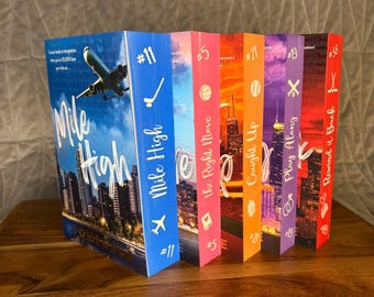May include: A collection of five paperback books standing upright, featuring colorful spines with titles including Mile High, The Right Move, Caught Up, Play Along, and Rewind It Back, each marked with a unique number and a small icon.