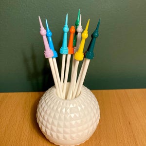 Puede incluir: Un recipiente blanco, esférico y texturizado que contiene una colección de palillos de cóctel coloridos. Los palillos tienen tallos blancos y están rematados con varios colores, incluyendo rosa, azul, naranja, amarillo y verde. El recipiente está sobre una superficie de madera.