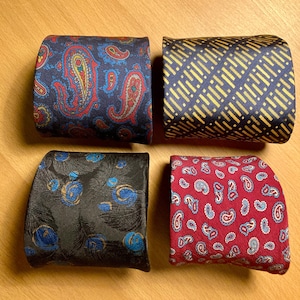 Könnte beinhalten: Vier gerollte Krawatten mit verschiedenen Mustern. Eine ist marineblau mit roten und gelben Paisley-Mustern, eine andere hat einen marineblauen Hintergrund mit goldenen diagonalen Linien. Die dritte ist schwarz mit blauen und goldenen federartigen Designs, und die letzte ist rot mit weißen und blauen Paisley-Mustern.